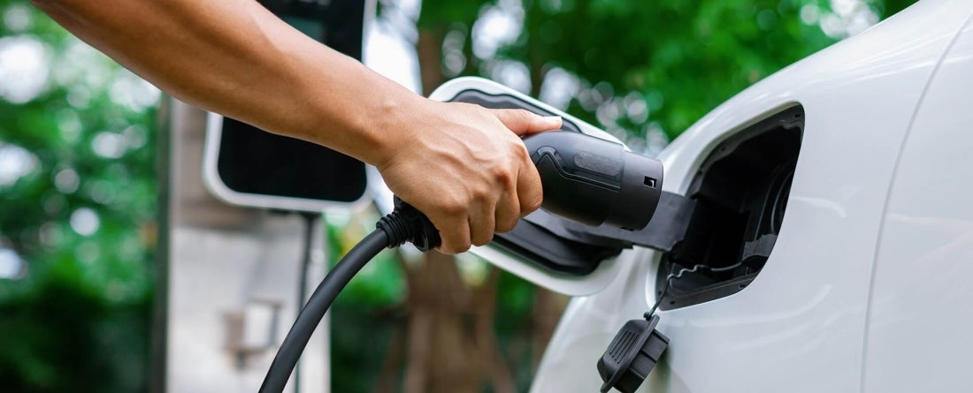 An arm holding an electric vehicle charger plugs the port into a white vehicle.