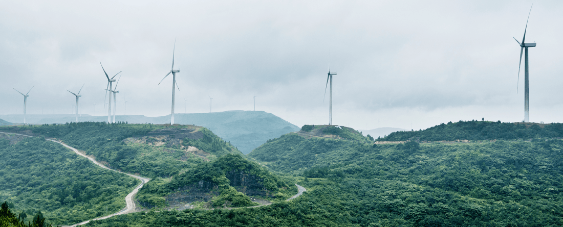 A group of wind turbines generate electricity on top of forested mountains with grey clouds in the sky.